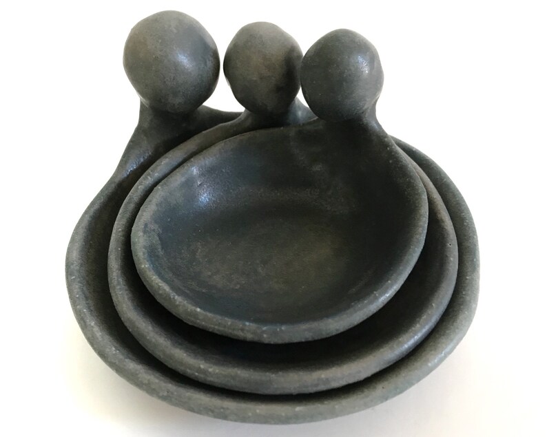 May include: Three grey ceramic nesting bowls with rounded handles. The bowls are stacked on top of each other.