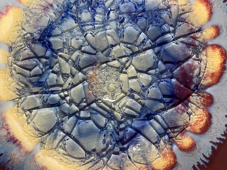 May include: Close-up of a ceramic plate with a crackled glaze in shades of blue, white, and yellow. The glaze has a textured, almost bubbly appearance.
