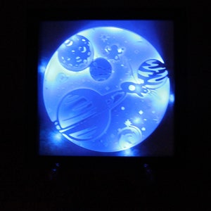 May include: A blue and white night light with a spaceship flying through space with planets and stars. The light is illuminated with white LED lights.