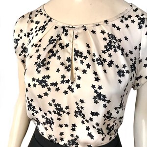 May include: A white silk blouse with a black star print and a keyhole neckline. The blouse has short sleeves and a relaxed fit.