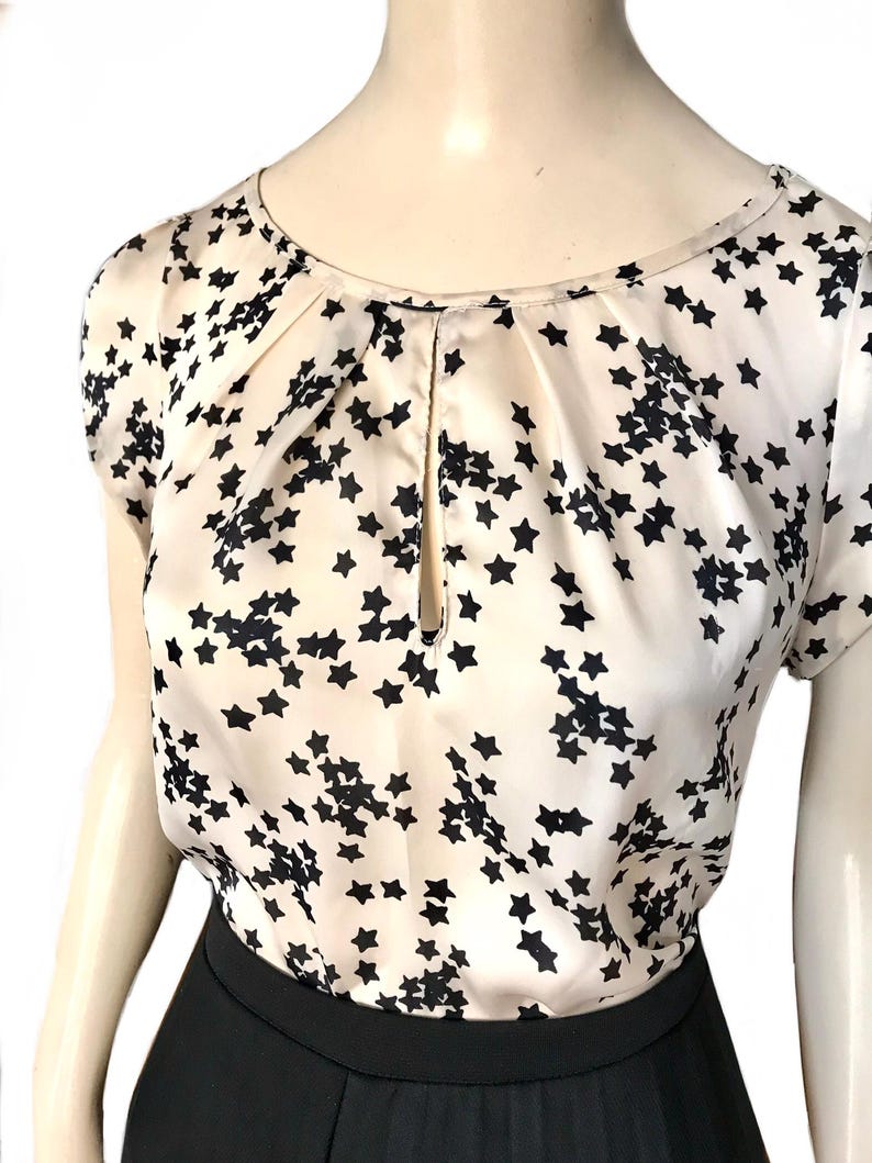 May include: A white silk blouse with a black star print and a keyhole neckline. The blouse has short sleeves and a relaxed fit.