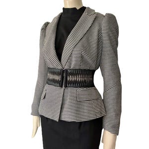 May include: A black and white houndstooth blazer with structured shoulders and long sleeves, worn over a solid black high-neck top and a black skirt. A wide, black, pleated elasticized cinch belt with a central clasp is worn over the blazer.