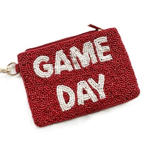 May include: A red beaded pouch with the words "GAME DAY" in white beads. The pouch has a zipper closure and a small tassel attached to the side. The background is white.