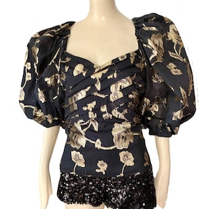 May include: A black top with gold floral embroidery and puffed sleeves is paired with black sequined shorts. The top features a sweetheart neckline and pleated detailing. The outfit is displayed on a mannequin.