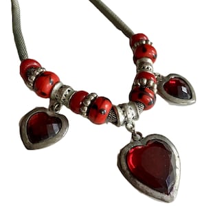 May include: A necklace featuring a light green cord strung with red beads that have black speckles, interspersed with silver-toned metal spacers. Three faceted dark red heart-shaped pendants with silver-toned metal frames hang from the necklace.