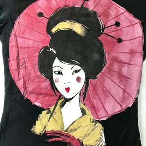 May include: Black t-shirt featuring a stylized illustration of a geisha. The geisha has black hair, red lips, and rosy cheeks, with a pink parasol in the background. She is wearing a yellow kimono and red gloves.