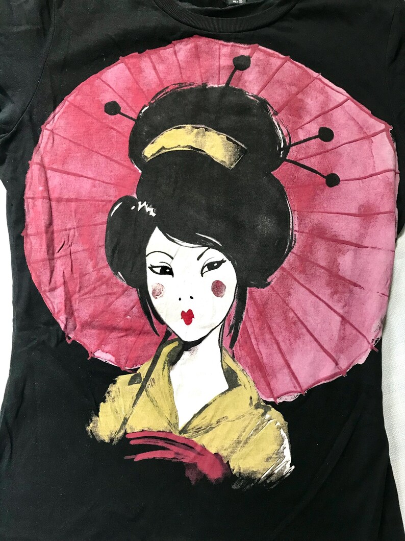 May include: Black t-shirt featuring a stylized illustration of a geisha. The geisha has black hair, red lips, and rosy cheeks, with a pink parasol in the background. She is wearing a yellow kimono and red gloves.