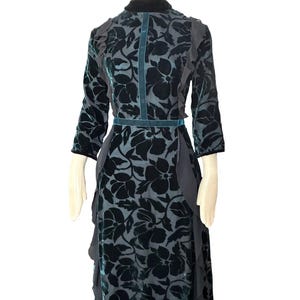 May include: A dark teal floral patterned dress with a high collar and three-quarter sleeves. The dress features a teal velvet belt and trim, with ruffled accents on the sleeves and skirt. The floral pattern is a darker shade of teal.