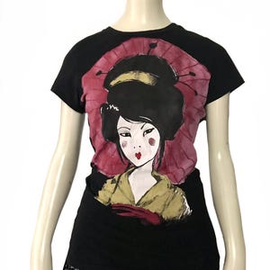 May include: Black t-shirt featuring a geisha illustration with a red parasol background. The geisha has black hair, red lips, and is wearing a yellow kimono. The bottom of the shirt has a black sequin trim.