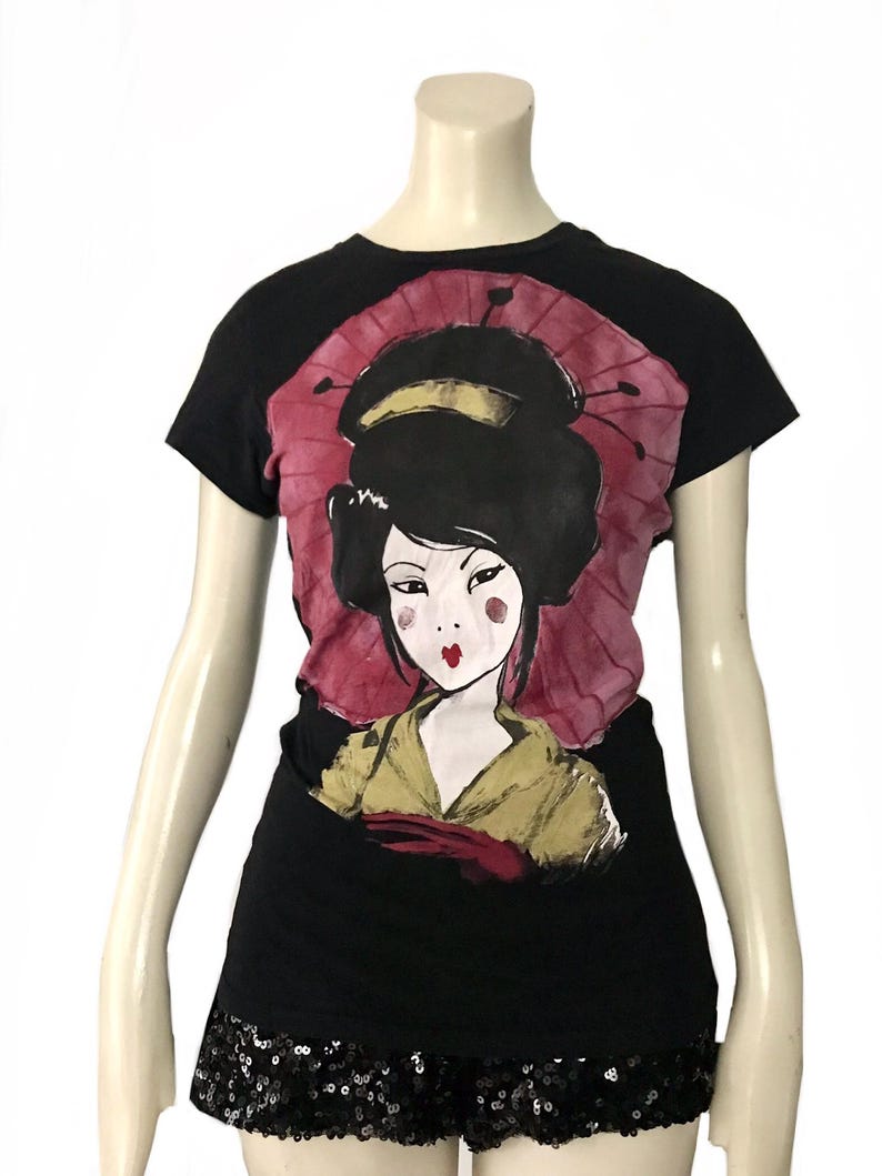 May include: Black t-shirt featuring a geisha illustration with a red parasol background. The geisha has black hair, red lips, and is wearing a yellow kimono. The bottom of the shirt has a black sequin trim.