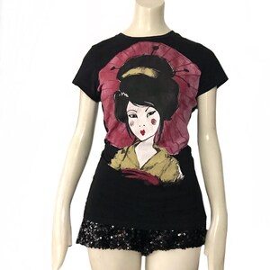 May include: Black t-shirt featuring a geisha illustration with a pink parasol. The geisha has black hair, red lips, and is wearing a yellow and red kimono. The shirt is paired with black sequin shorts, creating a stylish ensemble.
