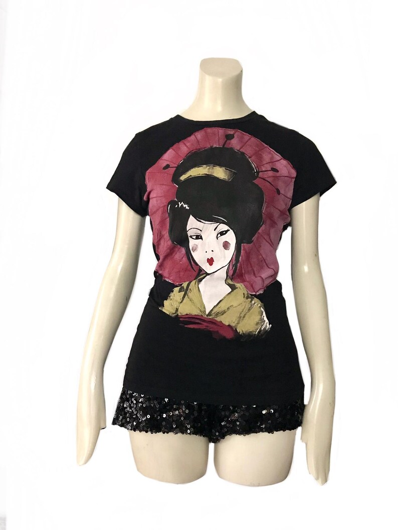 May include: Black t-shirt featuring a geisha illustration with a pink parasol. The geisha has black hair, red lips, and is wearing a yellow and red kimono. The shirt is paired with black sequin shorts, creating a stylish ensemble.