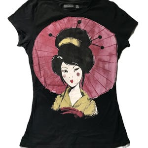 May include: Black t-shirt featuring a graphic of a geisha with a pink parasol. The geisha is depicted with black hair, red lips, and a yellow kimono. The design is centered on the front of the shirt.