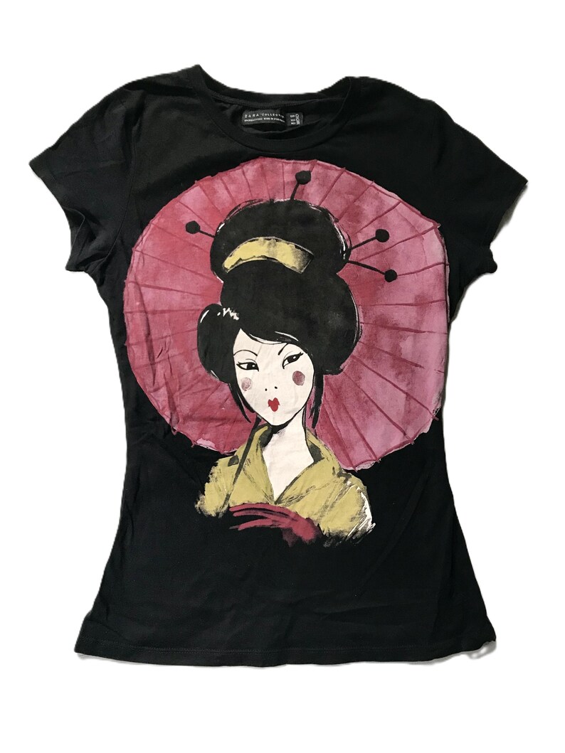 May include: Black t-shirt featuring a graphic of a geisha with a pink parasol. The geisha is depicted with black hair, red lips, and a yellow kimono. The design is centered on the front of the shirt.