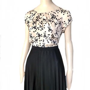 May include: A beige silk blouse with a black star print and a keyhole neckline. The blouse has short sleeves and is paired with a black pleated skirt.