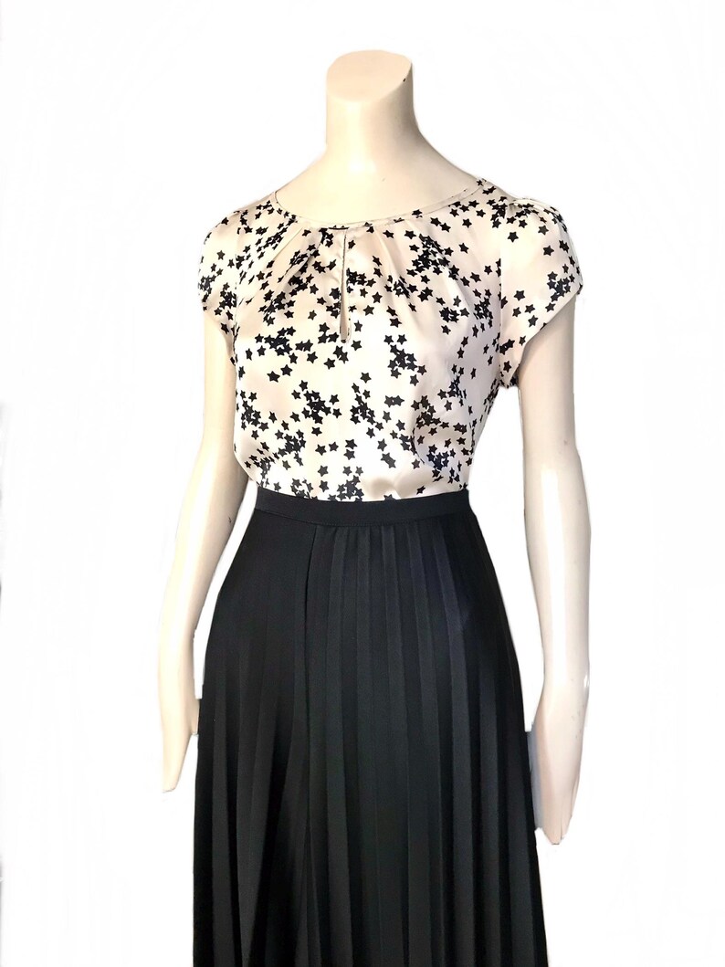 May include: A beige silk blouse with a black star print and a keyhole neckline. The blouse has short sleeves and is paired with a black pleated skirt.
