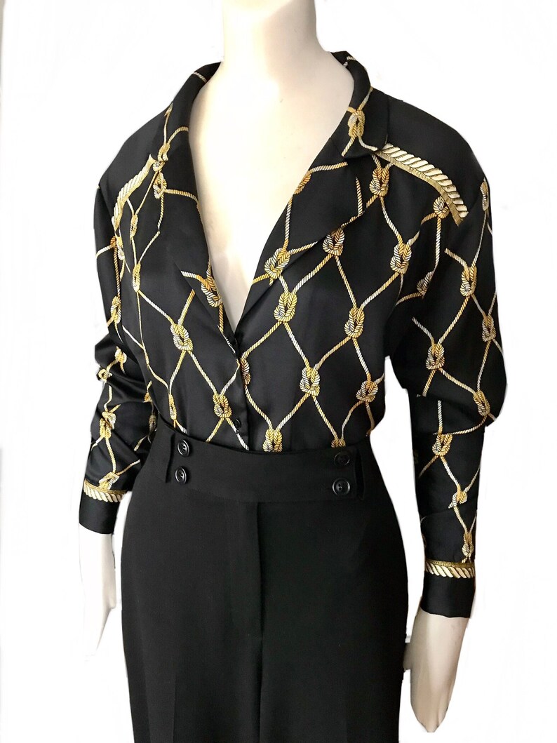 May include: A black and gold silk blouse with a nautical rope pattern. The blouse has a V-neckline, long sleeves, and a button-up front. The blouse is paired with black trousers.
