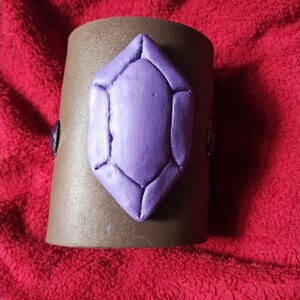 May include: A brown leather cuff bracelet with a large purple gem-shaped embellishment.