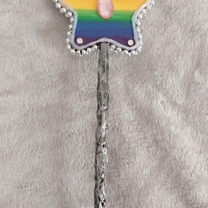May include: A rainbow-colored star-shaped wand with a silver-colored stick and a pink gem in the center. The star is decorated with silver beads.