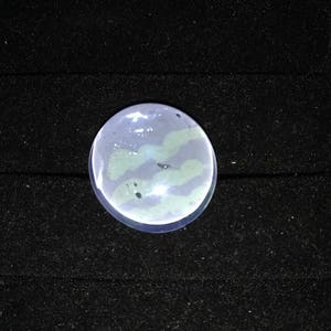 May include: A round, clear glass cabochon with a swirling, light blue and green pattern. The cabochon is set against a black background.