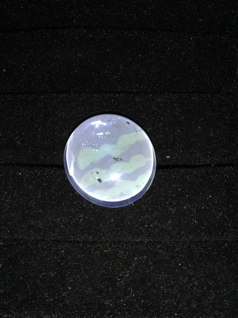May include: A round, clear glass cabochon with a swirling, light blue and green pattern. The cabochon is set against a black background.