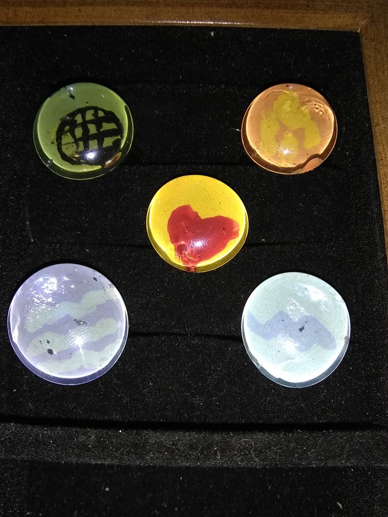 May include: Five round glass marbles with different designs. The marbles are displayed in a black velvet box. One marble is green with a black grid pattern, one is yellow with a red heart, one is orange with a brown swirl, and two are blue with a white swirl.
