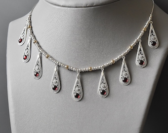 May include: Silver necklace with a delicate chain and multiple teardrop pendants. Each pendant features a filigree design and a red gemstone.