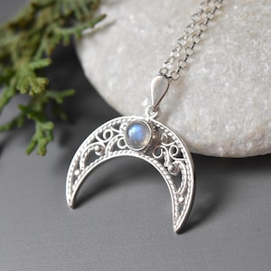 Könnte beinhalten: Halskette mit silbernem Halbmond-Anhänger und einem kleinen, runden, blauen Edelstein in der Mitte. Der Mond hat ein kompliziertes, offenes Design. Die Halskette ist filigran und silberfarben.