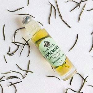 May include: A clear glass roller bottle with a green label that says "VISIONS Co. ROSEMARY ESSENTIAL OIL ROLLER SHOPVISIONS.COM 10 ML". The bottle is surrounded by dried tea leaves on a white background.