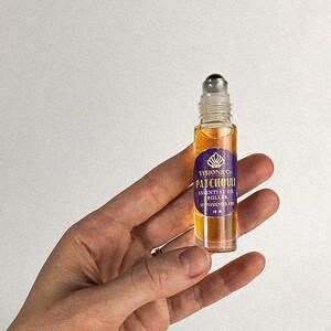 May include: A small glass bottle with a rollerball applicator filled with a light brown liquid. The bottle has a purple label with gold lettering that reads "VISIONS CO. PATCHOULI ESSENTIAL OIL ROLLER 10 ML".