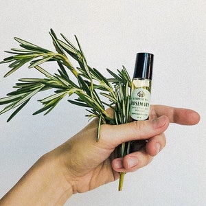 May include: A hand holding a small bottle of rosemary essential oil with a green label that says "VISION PURE ROSEMARY ESSENTIAL OIL".
