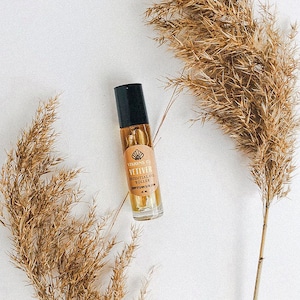 Puede incluir: Un frasco de vidrio con rodillo, tapa negra y una etiqueta que dice "VISIONS CO. VETIVER ESSENTIAL OIL ROLLER SOOTHING SERUM 10 ML". El frasco está sobre una superficie blanca con hierba de la pampa seca a cada lado.