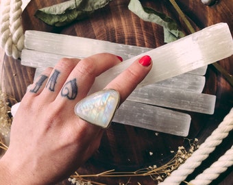 May include: A hand holding a selenite wand with a rainbow moonstone ring on the index finger. The wand is on a wooden tray with dried flowers and white rope.