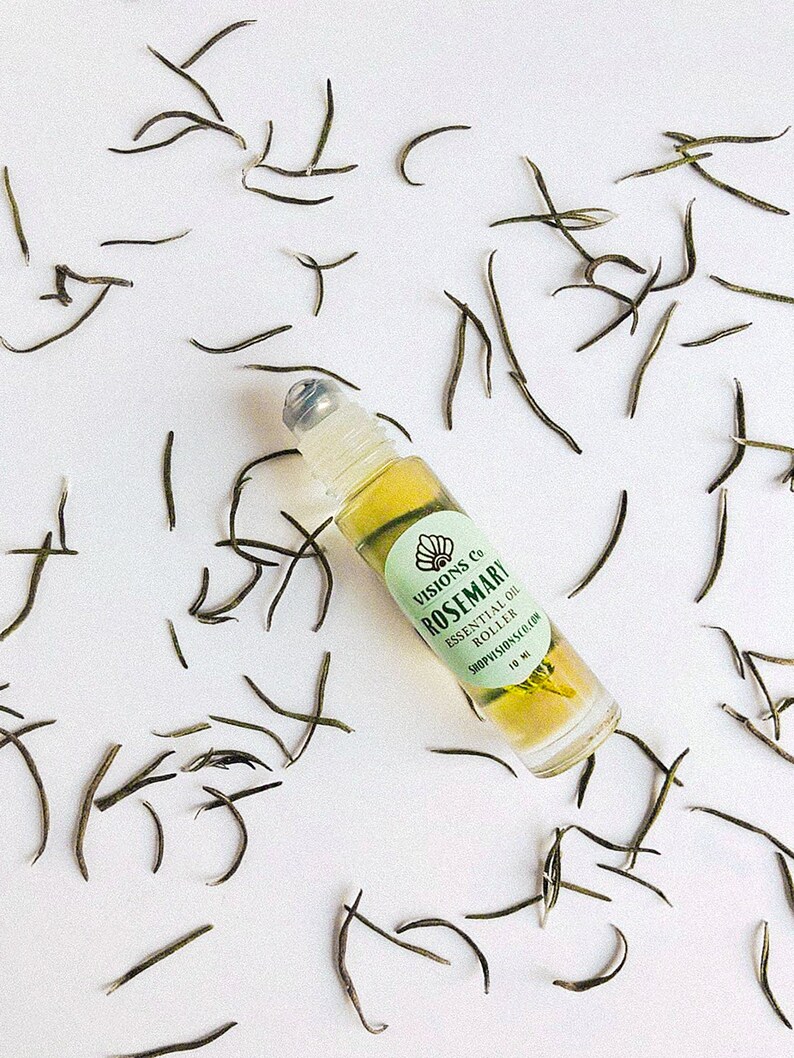 May include: A clear glass roller bottle with a green label that says "VISIONS Co. ROSEMARY ESSENTIAL OIL ROLLER SHOPVISIONS.COM 10 ML". The bottle is surrounded by dried tea leaves on a white background.