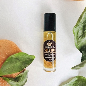 May include: A small glass bottle with a black cap and a gold label. The label reads "VISIONS Co. SOLSTICE PATCHOULI AND ORANGE ESSENTIAL OIL ROLLER SHOPVISIONS.COM 10 ML".