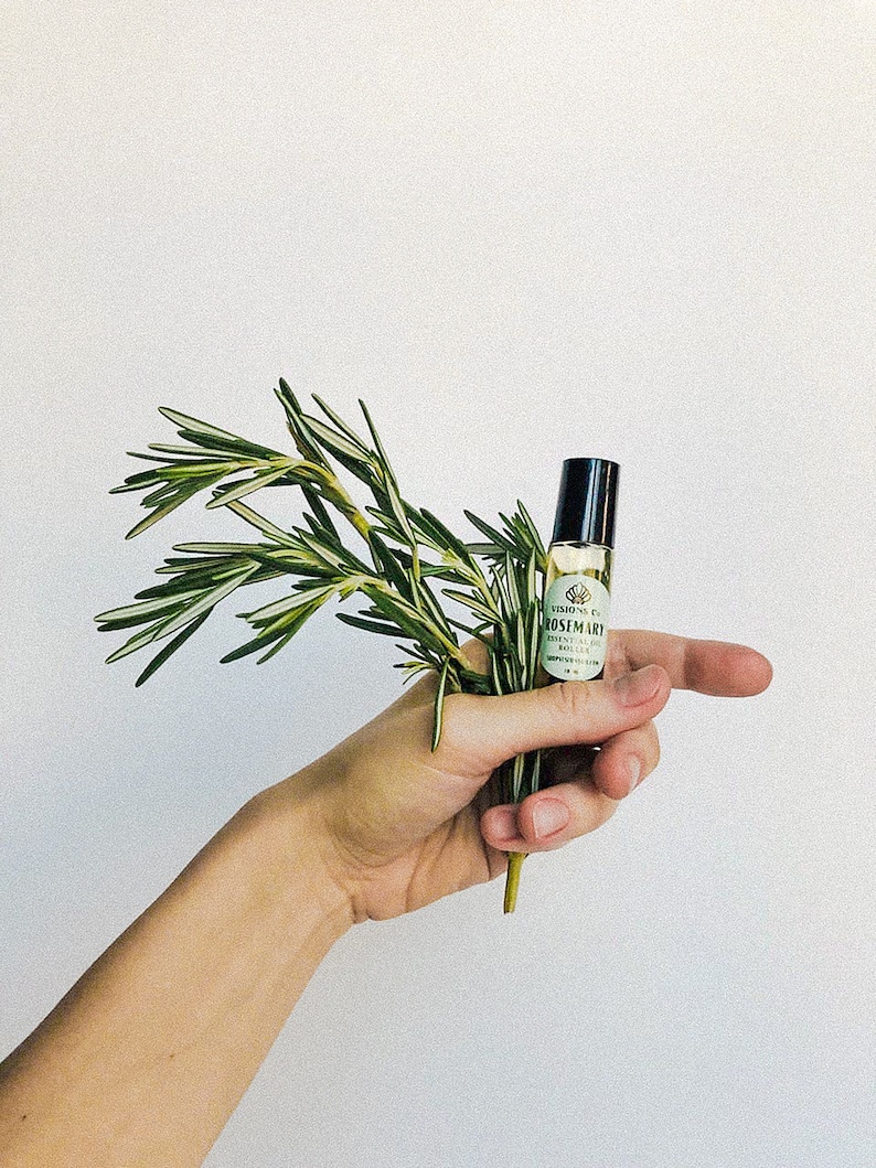 May include: A hand holding a small bottle of rosemary essential oil with a green label that says "VISION PURE ROSEMARY ESSENTIAL OIL".