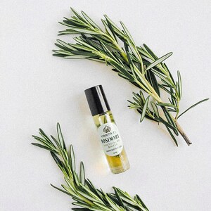 May include: A small glass bottle with a black rollerball top filled with a light yellow liquid. The bottle has a white label with the text "VISIONS CO. ROSEMARY ESSENTIAL OIL ROLLER" and a small graphic of a seashell. Two sprigs of rosemary are on either side of the bottle.