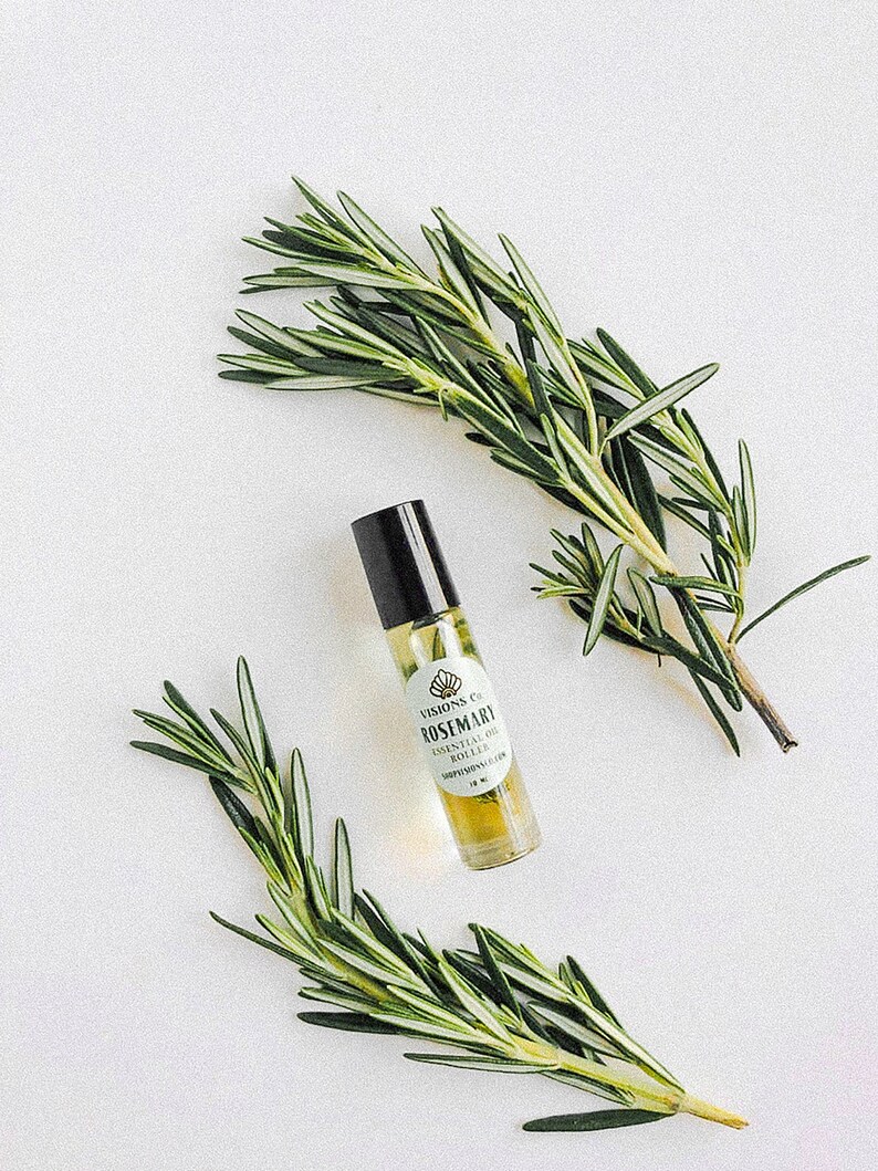 May include: A small glass bottle with a black rollerball top filled with a light yellow liquid. The bottle has a white label with the text "VISIONS CO. ROSEMARY ESSENTIAL OIL ROLLER" and a small graphic of a seashell. Two sprigs of rosemary are on either side of the bottle.