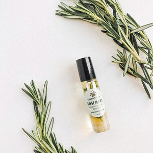 May include: A glass bottle of rosemary essential oil with a black roller ball top. The bottle has a white label with the text "VISIONS CO. ROSEMARY ESSENTIAL OIL ROLLER 100% NATURAL" and a small image of a seashell. Two sprigs of rosemary are on either side of the bottle.