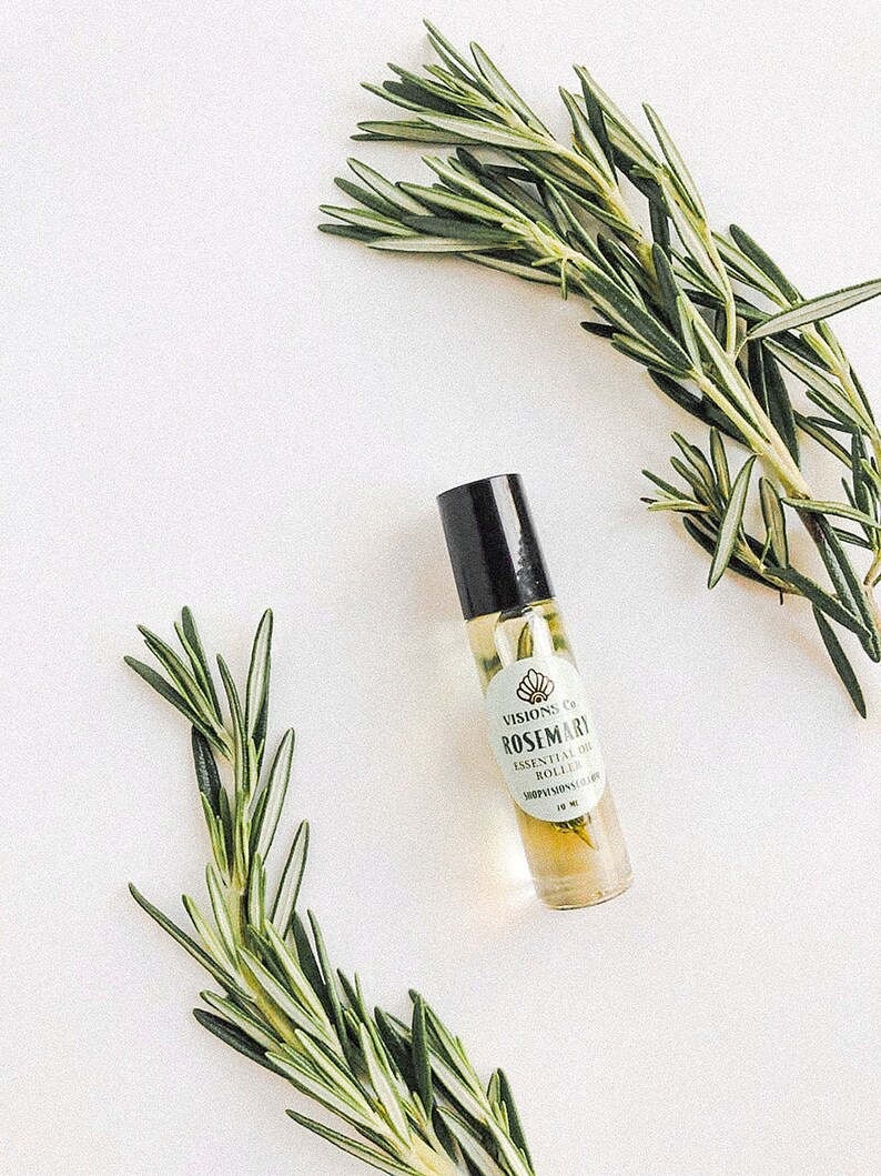 May include: A glass bottle of rosemary essential oil with a black roller ball top. The bottle has a white label with the text "VISIONS CO. ROSEMARY ESSENTIAL OIL ROLLER 100% NATURAL" and a small image of a seashell. Two sprigs of rosemary are on either side of the bottle.