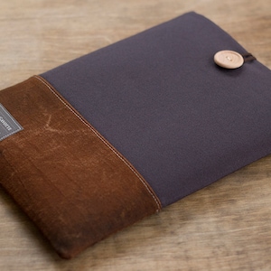 Peut inclure: Une pochette pour ordinateur portable de couleur gris foncé et marron avec un rabat en cuir marron et un bouton en bois. La pochette porte une étiquette sur laquelle est inscrit "POCHETTE GARÇONNE" avec le texte "Handmade in France" et "EST. 2014".