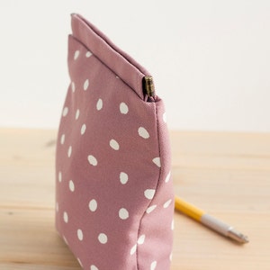 May include: A mauve-colored, polka-dotted pouch with a rectangular shape. The pouch is made of fabric and features white polka dots. A pencil is partially visible, resting on a wooden surface, suggesting its use for storing stationery or small items.