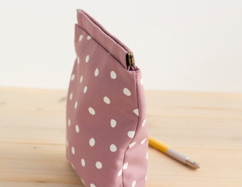 May include: A mauve-colored, polka-dotted pouch with a rectangular shape. The pouch is made of fabric and features white polka dots. A pencil is partially visible, resting on a wooden surface, suggesting its use for storing stationery or small items.