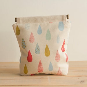 May include: A white canvas pouch with a zipper closure. The pouch is decorated with a repeating pattern of colourful raindrops.