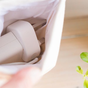 May include: A white charging adapter and cable are partially visible inside a small, off-white canvas pouch. The pouch is open, revealing the contents. A sprig of green leaves is in the background.