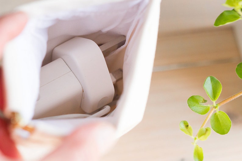 May include: A white charging adapter and cable are partially visible inside a small, off-white canvas pouch. The pouch is open, revealing the contents. A sprig of green leaves is in the background.