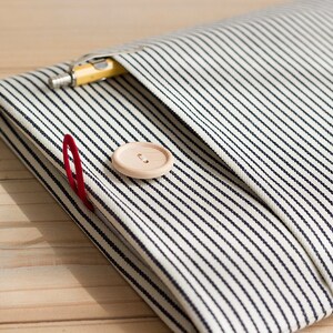 May include: A folded fabric item with a black and white striped pattern. A yellow pencil is tucked into a pocket. A red paper clip and a wooden button are also visible. The item is resting on a light wooden surface.