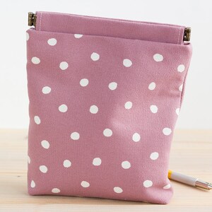 May include: A mauve-colored pouch with white polka dots. The pouch is rectangular with a flap and a metal clasp. A pen rests on the wooden surface next to the pouch. The pouch is made of fabric.