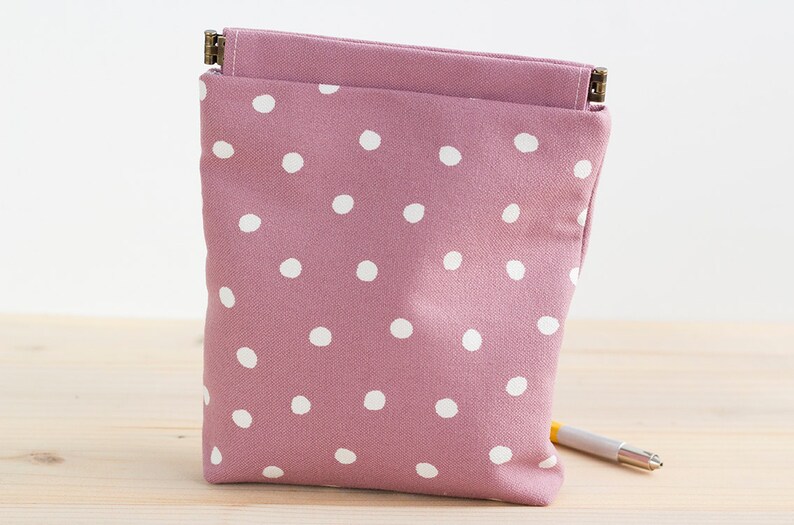 May include: A mauve-colored pouch with white polka dots. The pouch is rectangular with a flap and a metal clasp. A pen rests on the wooden surface next to the pouch. The pouch is made of fabric.