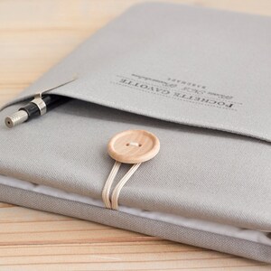 May include: A grey fabric notebook cover with a wooden button closure and a pen in the pocket. The cover has the text "POCHETTE CAYOTTE" printed on it.
